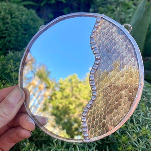 Può includere: Specchio decorativo rotondo con cornice color argento. Lo specchio riflette un cielo blu e alberi. Il lato destro presenta un design strutturato a scaglie in tonalità oro e marrone. Due piccoli anelli per appenderlo.