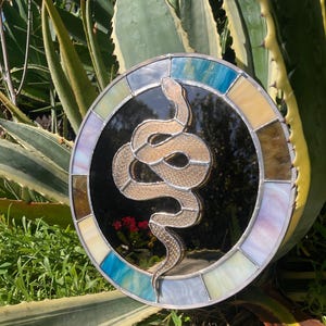 May include: A round stained glass piece featuring a coiled snake design. The snake is a light tan color, set against a black background. The border is composed of various colored glass pieces, including blue, yellow, and purple.