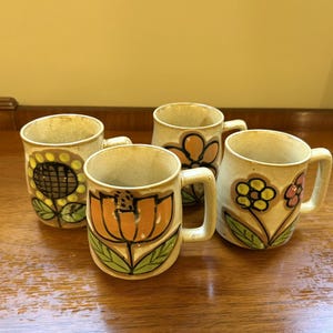 Peut inclure: Un ensemble de trois tasses en céramique avec une base de couleur crème et des motifs floraux peints à la main. Chaque tasse présente des motifs de fleurs distincts dans des tons orange, jaune et rose avec des feuilles vertes, soulignés de noir.