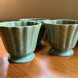 May include: Three sage green ceramic bowls with a fluted, scalloped rim design and a pedestal base. Each bowl features a mottled, speckled glaze finish with vertical ridges running along the exterior surface from the rim down to the base.