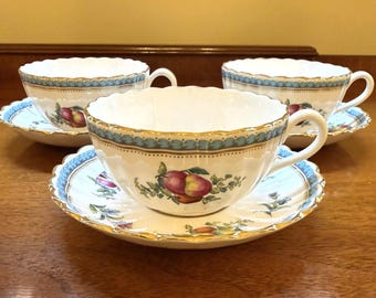 Puede incluir: Tres tazas de té de porcelana blanca con sus platos a juego, bordes festoneados y ribetes dorados. Cada pieza presenta un borde decorativo azul claro y una colorida ilustración de frutas, incluyendo manzanas rojas y amarillas con hojas verdes.