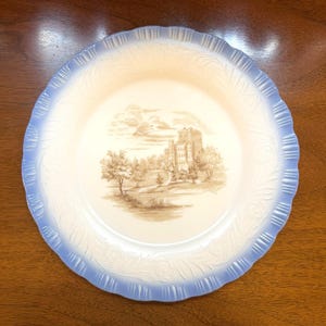May include: A round ceramic plate with a scalloped edge featuring a light blue gradient border and an embossed floral pattern. The centre of the plate displays a sepia-toned illustration of a stone castle surrounded by trees and a landscape.