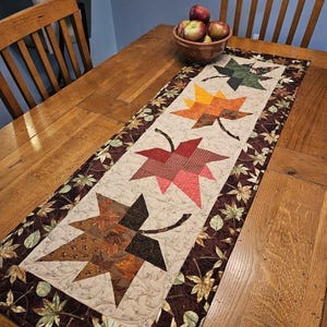 May include: A quilted table runner with a leaf design in green, orange, red, and brown. The runner has a beige background and a leaf border, placed on a wooden table. A bowl of apples is also visible.