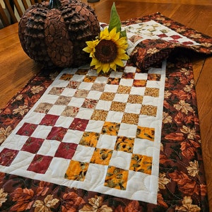 May include: A quilted table runner with a checkered pattern in shades of brown, white, and yellow. The runner has a border of brown fabric with a leaf pattern. A decorative pumpkin and a sunflower are on the table.