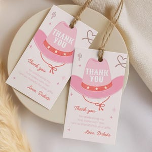 Op de afbeelding: Twee rechthoekige witte papieren cadeaulabels met jutekoord, elk voorzien van een roze cowboyhoedillustratie, steraccenten en de tekst: Thank you for celebrating my first rodeo with me! I am so thankful for your gifts. Love, Dakota.