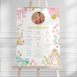 May include: A pink and gold decorative sign on an easel, celebrating a baby's first year. The sign includes a photo, floral and castle illustrations, and text with the baby's name, weight (20.9 lbs), height (29.5 inches), and other details.