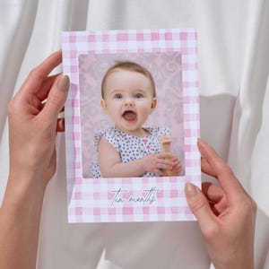 May include: A baby photo card with a pink and white gingham frame. The card features a smiling baby with blue eyes holding a wooden toy. The background is a soft pink floral pattern. The words 'Ten months' are written in cursive at the bottom.