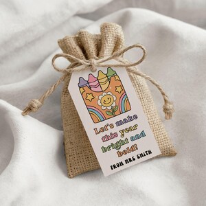 May include: A small burlap gift bag with a tag featuring a retro design of crayons, a smiling flower, and a rainbow. The tag reads "Let's make this year bright and bold! From Mrs Smith."