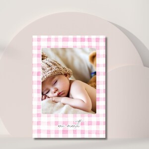 May include: A baby sleeping on a cream-colored surface, wearing a knit cap. The baby is the focal point, framed by a pink and white gingham pattern. The text 'one month' is written in cursive at the bottom of the frame. A teddy bear is partially visible in the background.