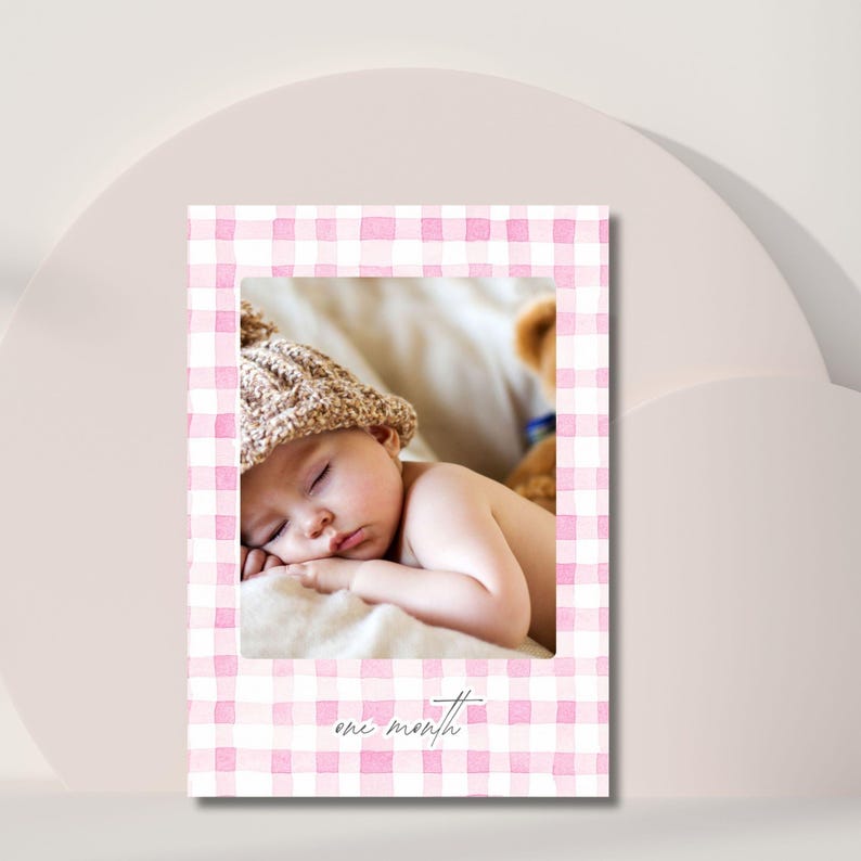 May include: A baby sleeping on a cream-colored surface, wearing a knit cap. The baby is the focal point, framed by a pink and white gingham pattern. The text 'one month' is written in cursive at the bottom of the frame. A teddy bear is partially visible in the background.