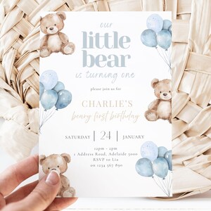 May include: A baby's first birthday invitation with a teddy bear theme. The invitation features the text "our little bear is turning one" and "Charlie's beary first birthday". It includes blue balloons and teddy bear illustrations.