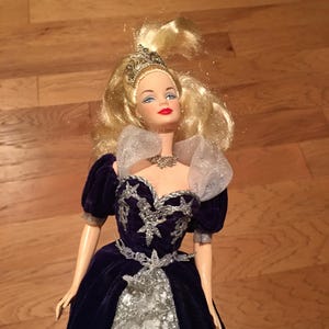 Puede incluir: Mu&ntilde;eca con cabello rubio y ojos azules, vestida con un vestido de terciopelo azul oscuro con adornos plateados. El vestido presenta un corpi&ntilde;o plateado y una falda blanca. La mu&ntilde;eca est&aacute; adornada con una tiara y un collar plateados.