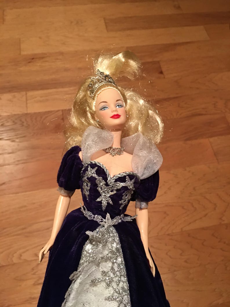 Puede incluir: Mu&ntilde;eca con cabello rubio y ojos azules, vestida con un vestido de terciopelo azul oscuro con adornos plateados. El vestido presenta un corpi&ntilde;o plateado y una falda blanca. La mu&ntilde;eca est&aacute; adornada con una tiara y un collar plateados.