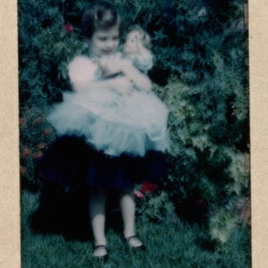 Puede incluir: Fotograf&iacute;a vintage de una ni&ntilde;a peque&ntilde;a sosteniendo una mu&ntilde;eca. La ni&ntilde;a lleva un vestido azul claro con una falda de tul blanca y una falda azul oscuro. La ni&ntilde;a est&aacute; de pie sobre hierba verde frente a un arbusto verde.