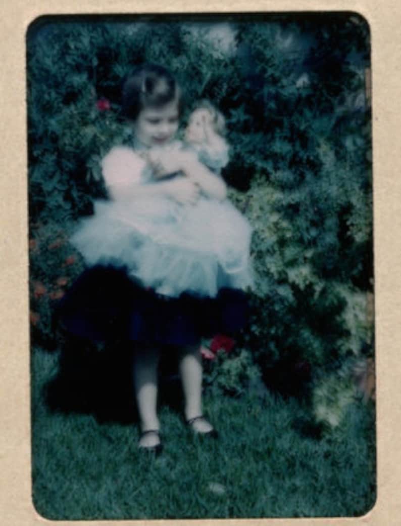 Puede incluir: Fotograf&iacute;a vintage de una ni&ntilde;a peque&ntilde;a sosteniendo una mu&ntilde;eca. La ni&ntilde;a lleva un vestido azul claro con una falda de tul blanca y una falda azul oscuro. La ni&ntilde;a est&aacute; de pie sobre hierba verde frente a un arbusto verde.