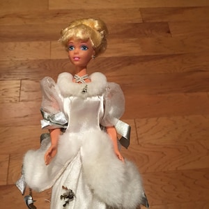 Puede incluir: Una mu&ntilde;eca con un vestido de sat&eacute;n blanco con adornos de piel sint&eacute;tica y detalles plateados. La mu&ntilde;eca tiene el pelo rubio recogido, ojos azules y un collar. El vestido presenta un cuello de piel sint&eacute;tica y un lazo decorativo.