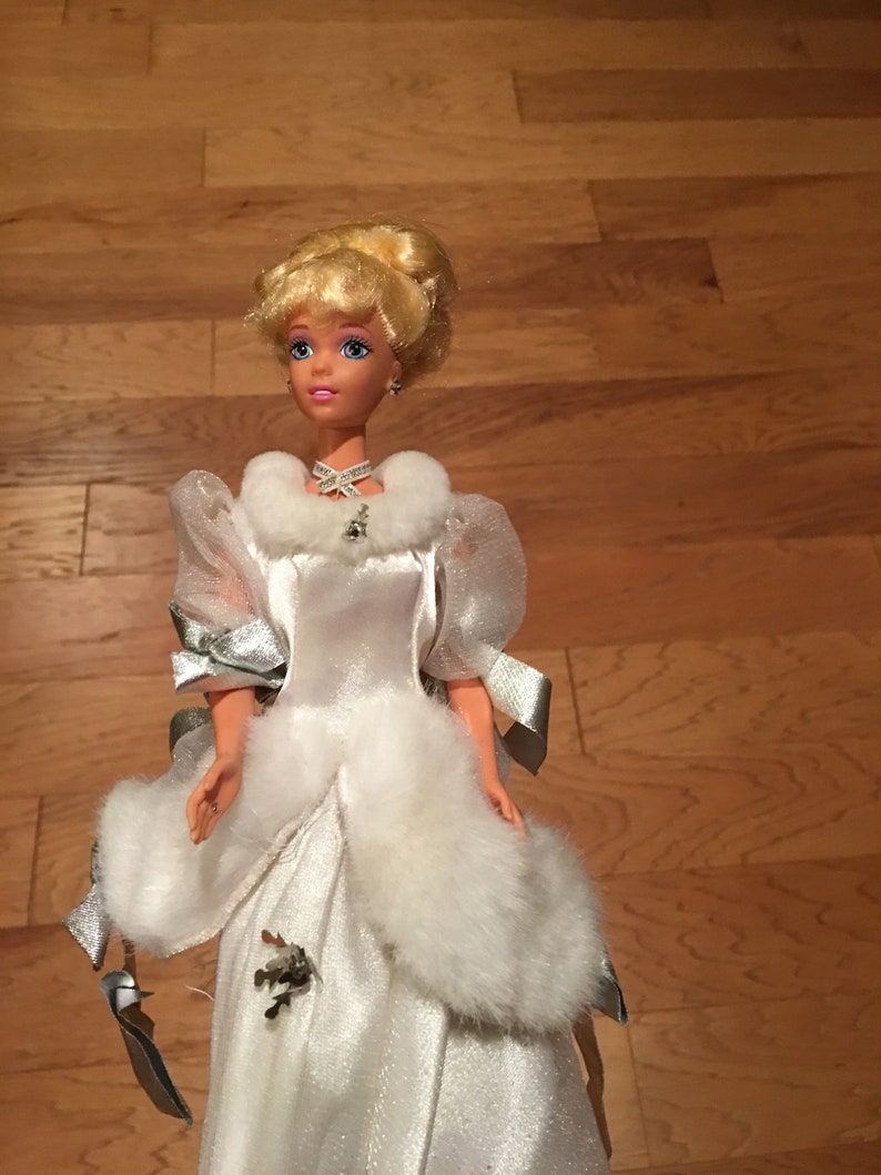 Puede incluir: Una mu&ntilde;eca con un vestido de sat&eacute;n blanco con adornos de piel sint&eacute;tica y detalles plateados. La mu&ntilde;eca tiene el pelo rubio recogido, ojos azules y un collar. El vestido presenta un cuello de piel sint&eacute;tica y un lazo decorativo.