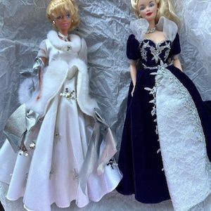 Puede incluir: Se muestran dos mu&ntilde;ecas vintage, cada una con un vestido elegante. Una mu&ntilde;eca lleva un vestido blanco con detalles plateados y una capa con ribete de piel. La otra mu&ntilde;eca lleva un vestido de terciopelo azul oscuro con una falda de encaje blanco y una tiara.