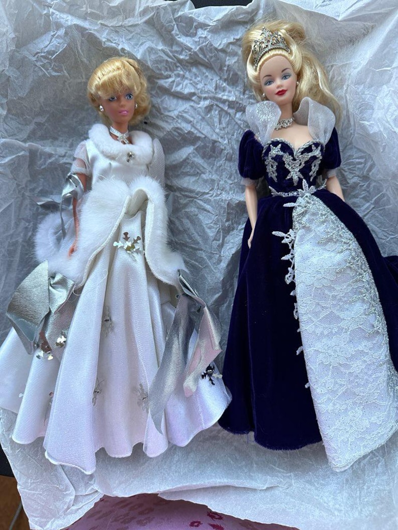 Puede incluir: Se muestran dos mu&ntilde;ecas vintage, cada una con un vestido elegante. Una mu&ntilde;eca lleva un vestido blanco con detalles plateados y una capa con ribete de piel. La otra mu&ntilde;eca lleva un vestido de terciopelo azul oscuro con una falda de encaje blanco y una tiara.