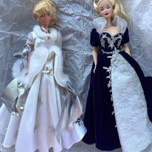 Puede incluir: Dos mu&ntilde;ecas rubias con vestidos elegantes. Una mu&ntilde;eca lleva un vestido blanco con detalles plateados y una estola de piel sint&eacute;tica. La otra mu&ntilde;eca viste un vestido de terciopelo azul oscuro con encaje blanco y una tiara. Ambas mu&ntilde;ecas tienen rasgos faciales detallados.