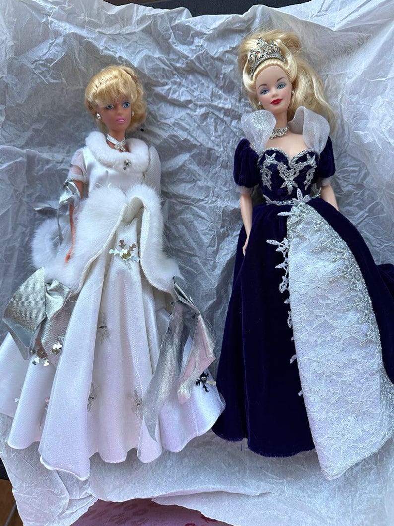 Puede incluir: Dos mu&ntilde;ecas rubias con vestidos elegantes. Una mu&ntilde;eca lleva un vestido blanco con detalles plateados y una estola de piel sint&eacute;tica. La otra mu&ntilde;eca viste un vestido de terciopelo azul oscuro con encaje blanco y una tiara. Ambas mu&ntilde;ecas tienen rasgos faciales detallados.