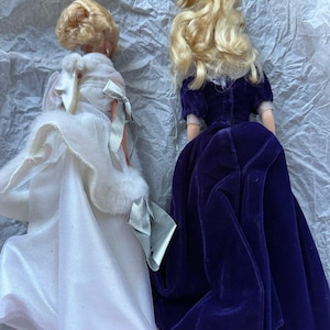 Puede incluir: Dos mu&ntilde;ecas de moda vintage con cabello rubio. Una mu&ntilde;eca viste un vestido de sat&eacute;n blanco con una estola de piel sint&eacute;tica y un lazo plateado. La otra mu&ntilde;eca lleva un vestido de terciopelo morado oscuro. Ambas mu&ntilde;ecas est&aacute;n de espaldas.