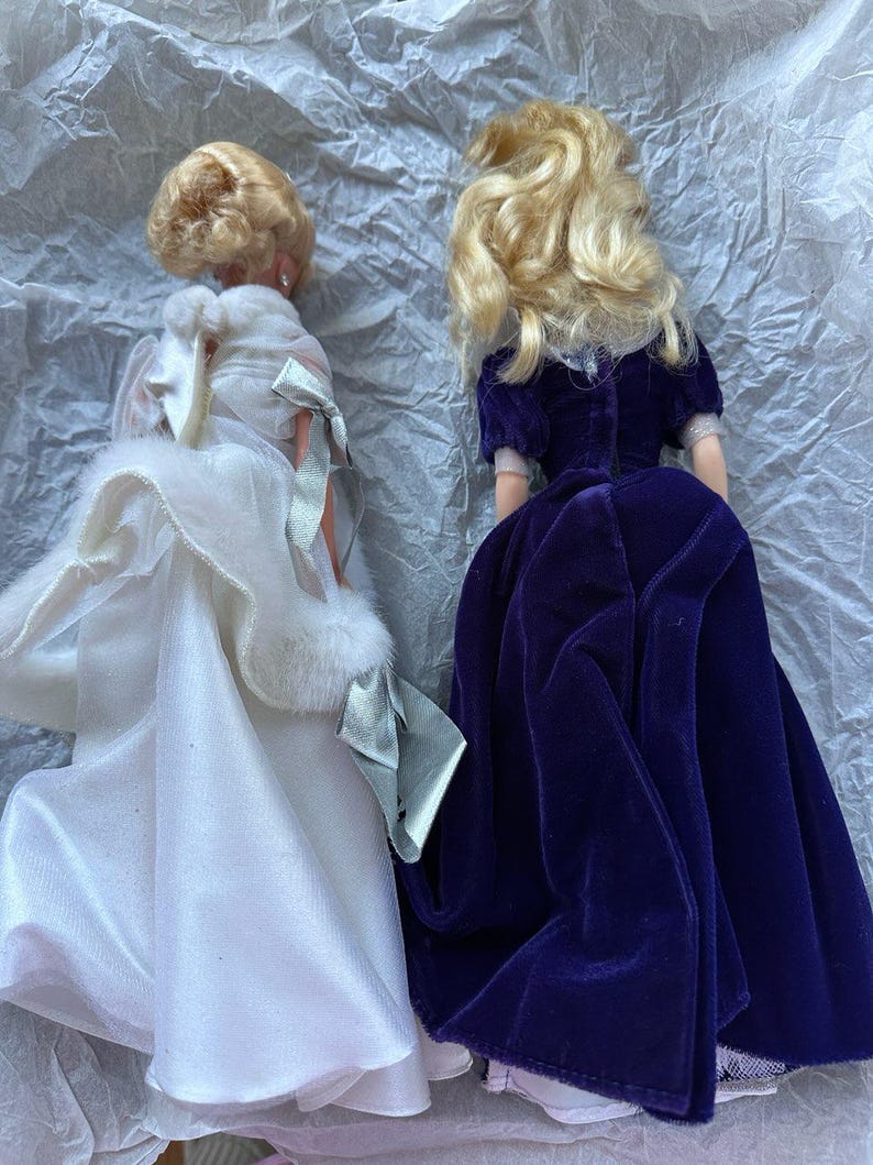 Puede incluir: Dos mu&ntilde;ecas de moda vintage con cabello rubio. Una mu&ntilde;eca viste un vestido de sat&eacute;n blanco con una estola de piel sint&eacute;tica y un lazo plateado. La otra mu&ntilde;eca lleva un vestido de terciopelo morado oscuro. Ambas mu&ntilde;ecas est&aacute;n de espaldas.