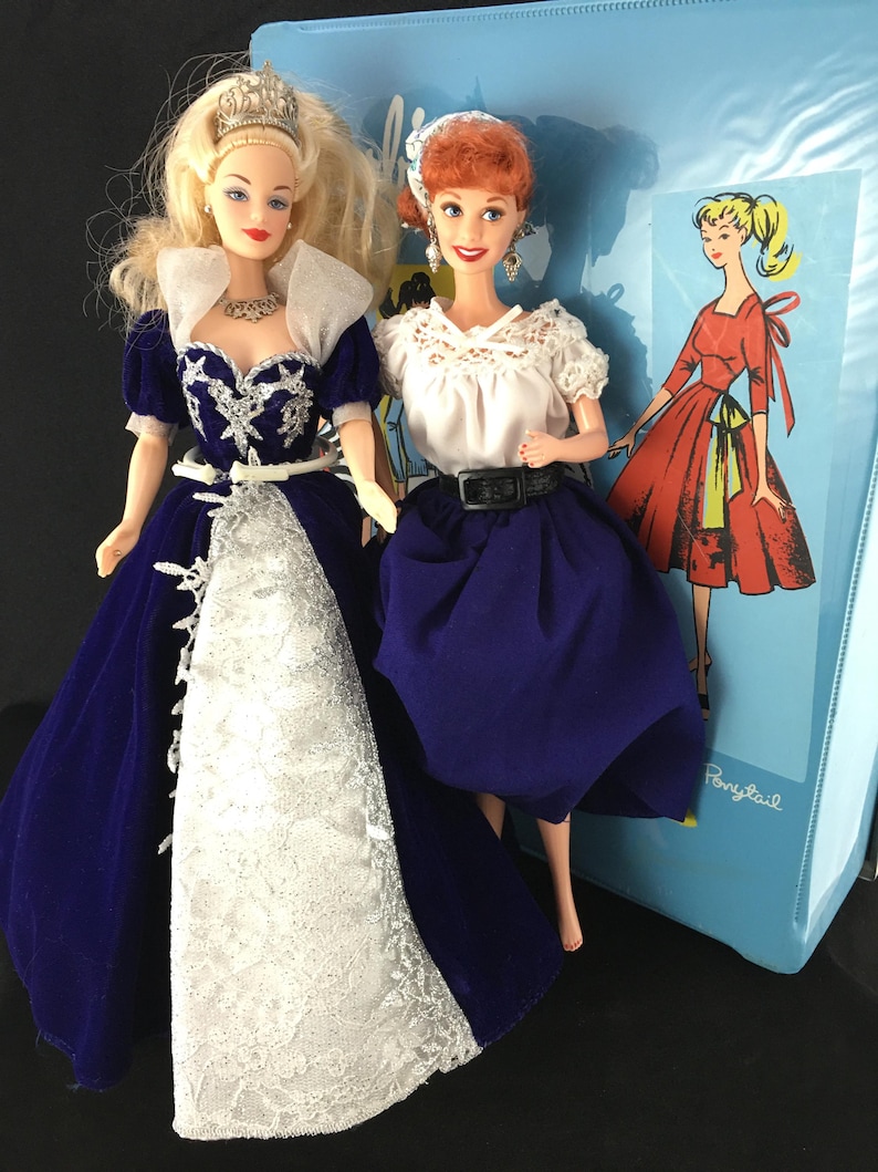 Puede incluir: Dos mu&ntilde;ecas vintage. Una mu&ntilde;eca viste un vestido de terciopelo azul con encaje blanco y una tiara plateada. La otra mu&ntilde;eca viste una blusa blanca, una falda azul y un cintur&oacute;n negro. Un archivador azul con un dibujo de una mujer con un vestido rojo est&aacute; en el fondo.