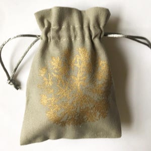 Peut inclure: Un petit sac à cordon de serrage vert sauge avec un motif floral doré. Le sac est fait d'un tissu texturé et possède une fermeture à cordon de serrage gris argenté. Le motif floral est centré sur le devant du sac.
