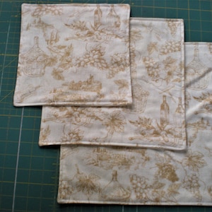 May include: Three white fabric napkins with a brown and white pattern of grapes and wine bottles. The napkins are stacked on top of each other, with the largest napkin on the bottom and the smallest napkin on top.