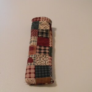 May include: A patchwork fabric tube with red, green, brown, and white squares and a heart design. The tube is made of cotton and is perfect for storing small items.