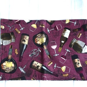 May include: A rectangular burgundy placemat with a repeating pattern of wine bottles, corkscrews, wine glasses, and corks. The wine bottles have labels with gold accents. The wine glasses are filled with pink and yellow wine.
