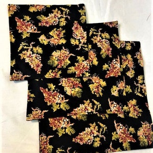 May include: Four black fabric placemats with a repeating pattern of grapes and leaves in shades of yellow, orange, and red. The grape clusters are detailed, and the leaves have a realistic appearance.
