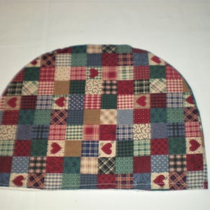 May include: A quilted tea cozy with a patchwork design. The cozy features a variety of square patterns in red, green, blue, and tan, with heart-shaped accents. The cozy is designed to fit over a teapot.