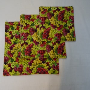 May include: Four fabric placemats with a pattern of green and red grapes. The placemats are stacked on top of each other.
