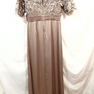 May include: A floor-length, taupe-colored dress. The bodice features a floral lace overlay with short sleeves. The waist is cinched with a pleated band, and the skirt flows smoothly. The dress is made of a satin-like material.