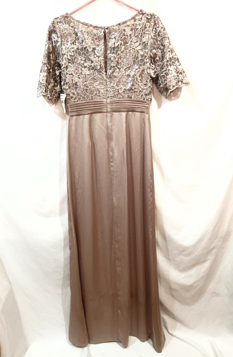 May include: A floor-length, taupe-colored dress. The bodice features a floral lace overlay with short sleeves. The waist is cinched with a pleated band, and the skirt flows smoothly. The dress is made of a satin-like material.