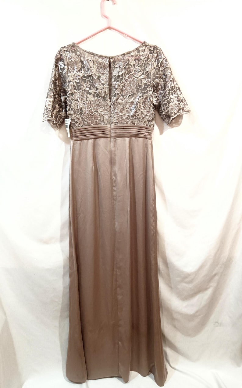 May include: A floor-length, taupe-colored dress with a lace bodice and short sleeves. The dress features a fitted waist and a flowing skirt. The back of the dress has a keyhole opening. The dress is hanging on a pink hanger.