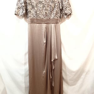May include: A floor-length, taupe-colored formal dress. The bodice features a floral lace overlay with short sleeves. The waist is cinched with a pleated band, and the skirt flows with a draped detail. The dress is made of a satin-like material.