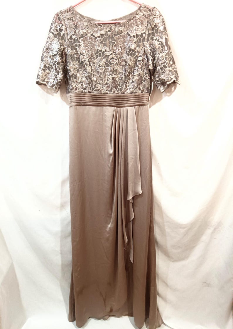 May include: A floor-length, taupe-colored formal dress. The bodice features a floral lace overlay with short sleeves. The waist is cinched with a pleated band, and the skirt flows with a draped detail. The dress is made of a satin-like material.