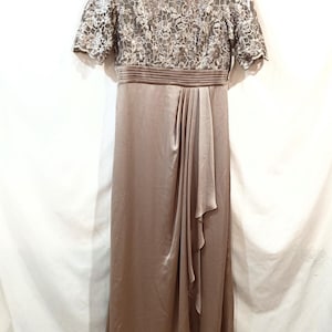 May include: A floor-length, taupe-colored formal dress. The bodice features a floral lace overlay with short sleeves. The waist is cinched with a pleated band, and the skirt has a draped, flowing design. The dress hangs on a pink hanger.