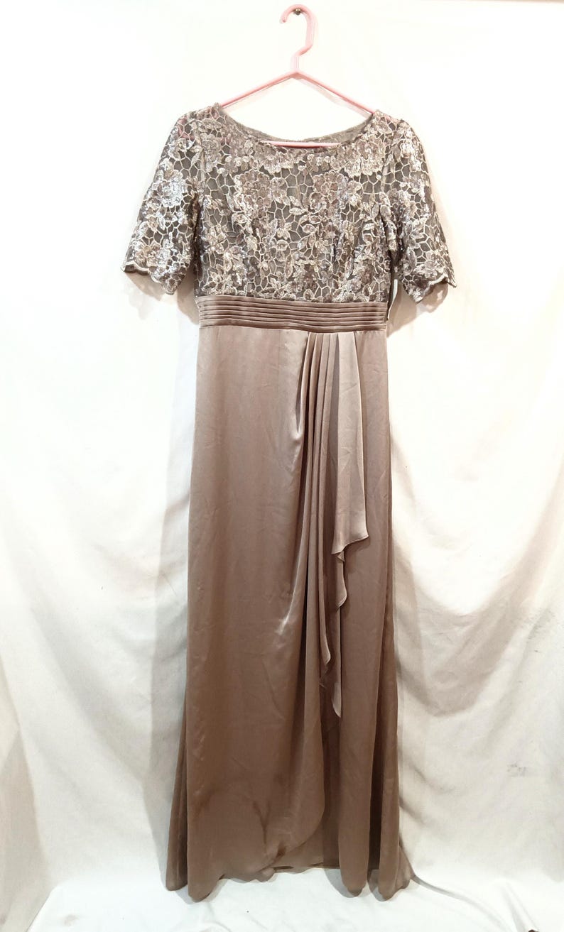 May include: A floor-length, taupe-colored formal dress. The bodice features a floral lace overlay with short sleeves. The waist is cinched with a pleated band, and the skirt has a draped, flowing design. The dress hangs on a pink hanger.
