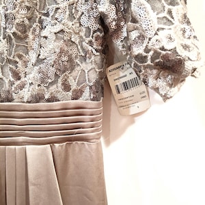 May include: A formal, floor-length dress in a neutral champagne color. The bodice features intricate sequined lace, transitioning to a pleated satin skirt. The dress has short sleeves and a defined waist. A tag with the brand name and style information is attached.