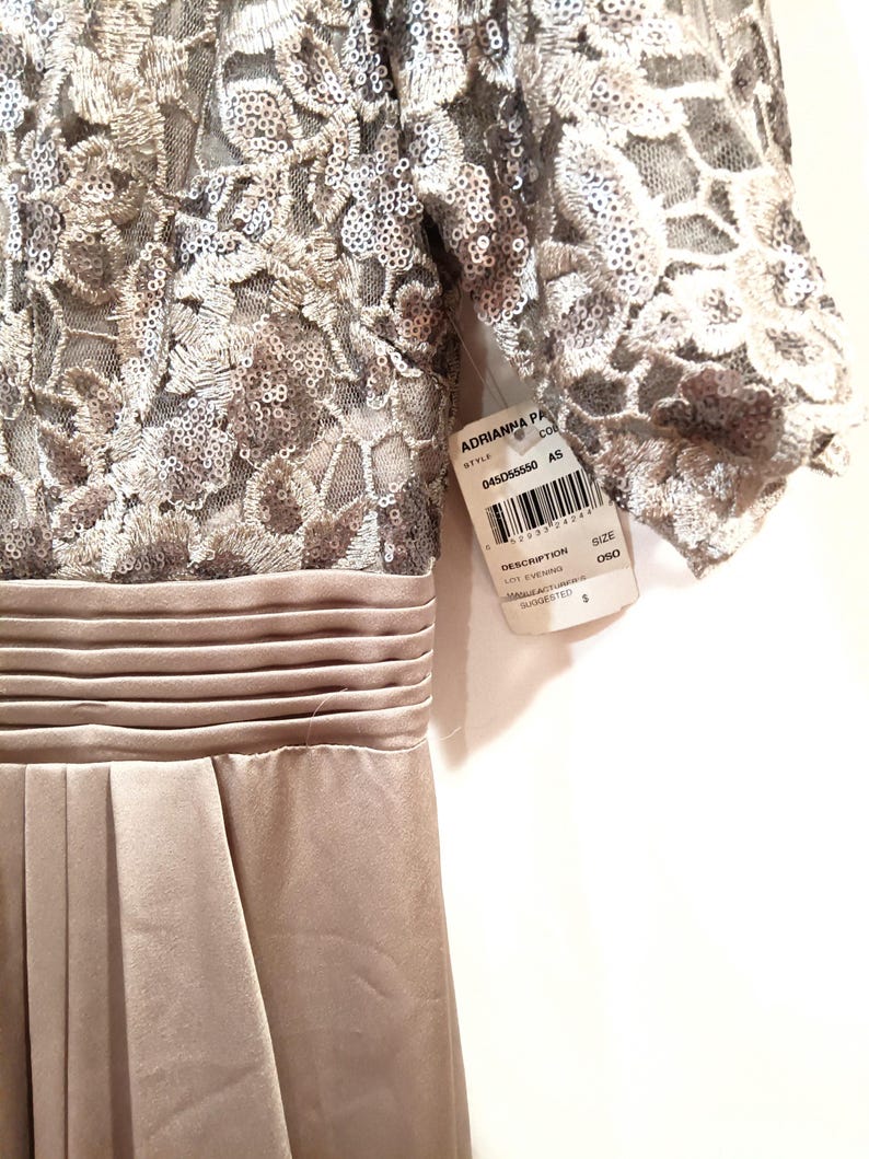 May include: A formal, floor-length dress in a neutral champagne color. The bodice features intricate sequined lace, transitioning to a pleated satin skirt. The dress has short sleeves and a defined waist. A tag with the brand name and style information is attached.