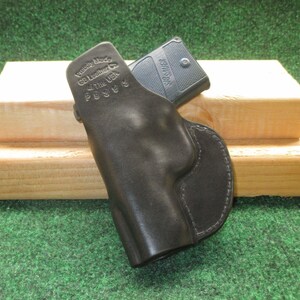 May include: A black leather holster with a firearm inside. The holster is stamped with "Proudly Made by GB Leather in the USA" and the model number P938S. The holster is resting on a wooden block against a green background.