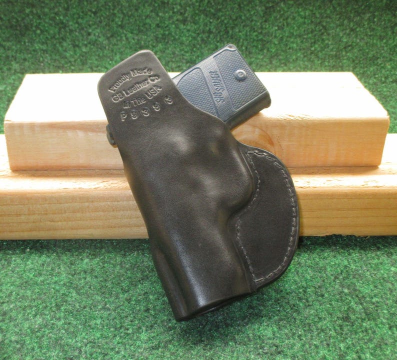 May include: A black leather holster with a firearm inside. The holster is stamped with "Proudly Made by GB Leather in the USA" and the model number P938S. The holster is resting on a wooden block against a green background.
