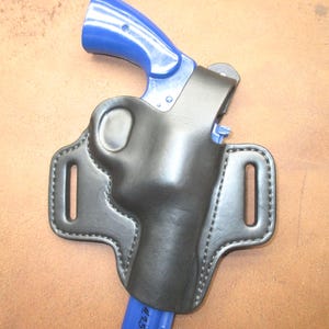 May include: A black leather holster with a blue handle. The holster has a stitched border and two slots for a belt. The background is a light brown color.