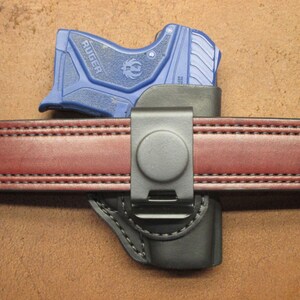 May include: A blue Ruger handgun in a black holster attached to a red leather belt with a metal clip. The belt is stitched with a double row of thread.