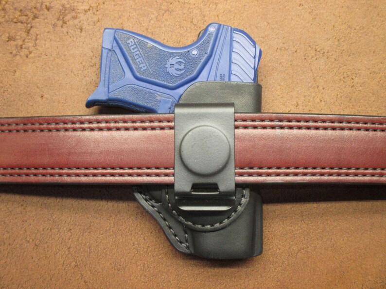 May include: A blue Ruger handgun in a black holster attached to a red leather belt with a metal clip. The belt is stitched with a double row of thread.