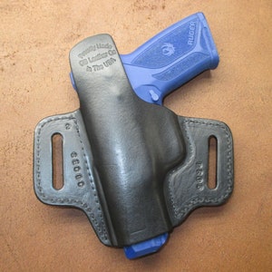 Może przedstawiać: Czarna sk&oacute;rzana kabura z niebieskim pistoletem Ruger w środku. Kabura ma pętlę na pasek i jest oznaczona napisem "Proudly Made by GG Leather Co. In The USA".